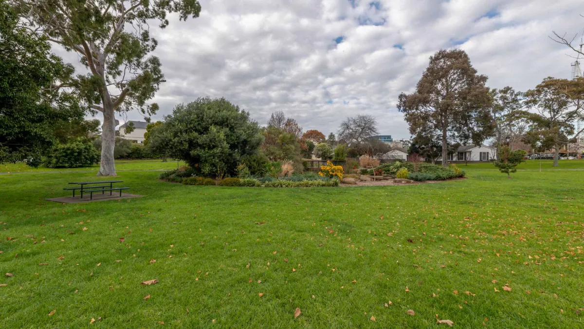 Round garden containing small plants and trees, in the middle of a large grass area. There is a table and two bench chairs under a tall tree to the left.