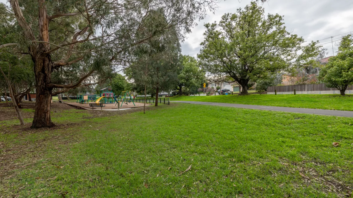 Small grass area with distant playground shaded by several tall trees. There is a walking path and fence to the right.