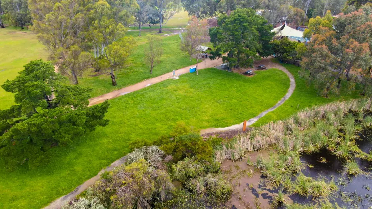 Aerial view of small grass area and walking path, on the edge of a wetland to the bottom right. There arev large scattered trees and a partly visible playground.