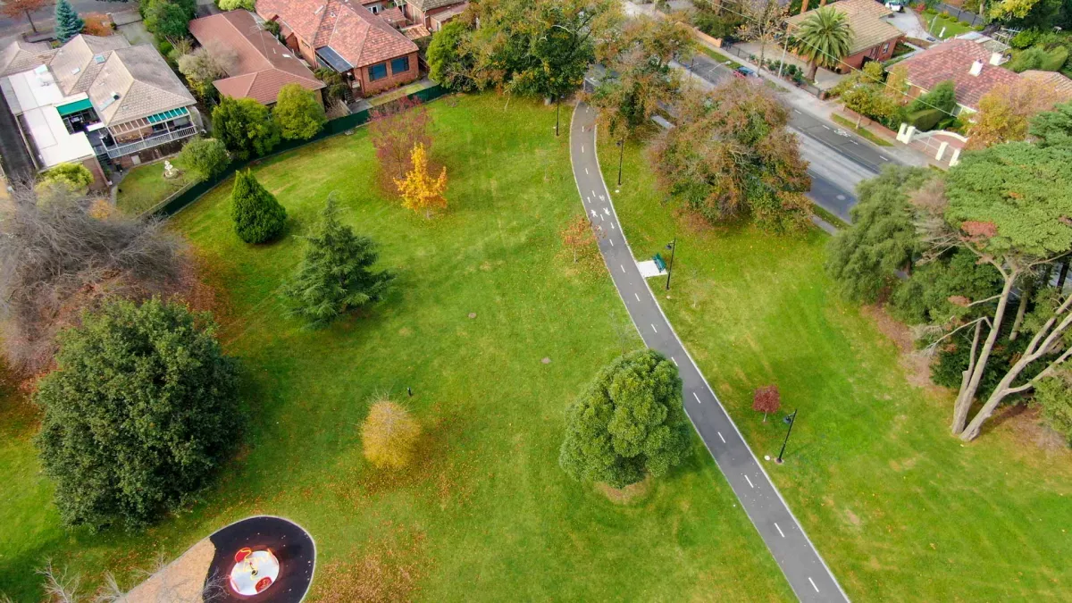 Aerial view of grass area with a footpath through the middle and scattered medium-sized trees. There is part of a playground to the bottom left and houses at the top.