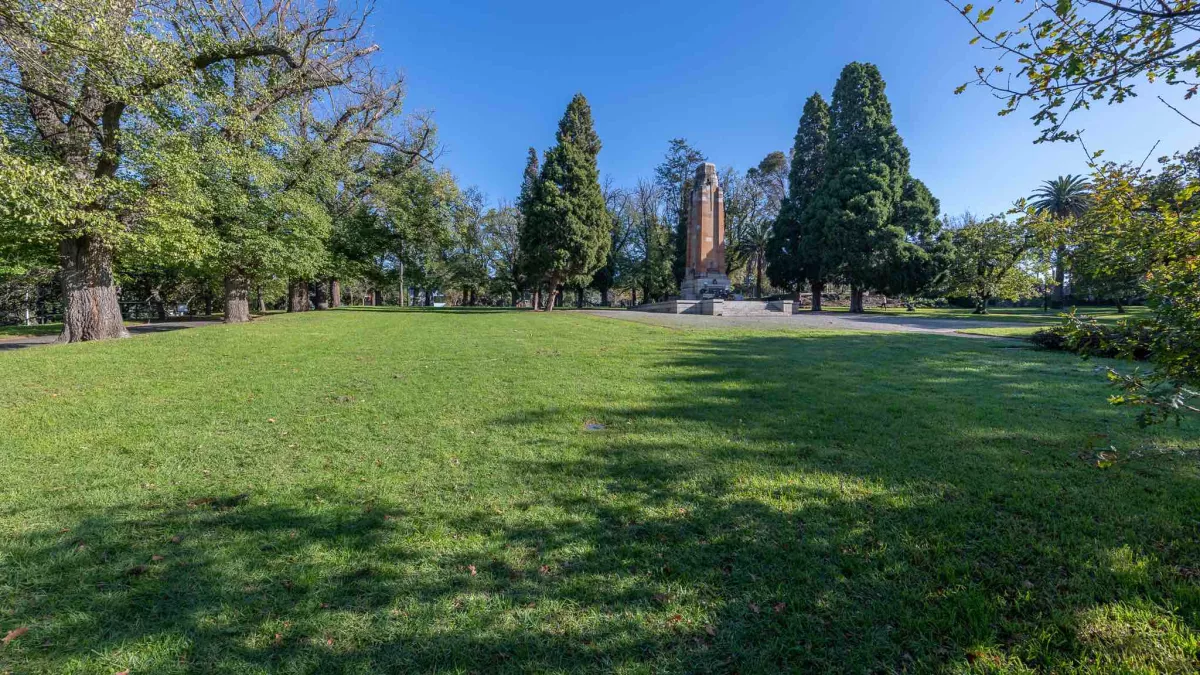 Area of lush grass in sunshine with concrete memorial structure and tall trees at the far end. There are shadows in the foreground.