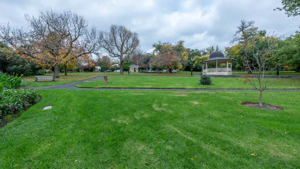 Large grass area with rotunda to the far right and a mix of bare and leafy trees. There is a park bench next to an L-shaped footpath and a small bare tree to the near right.