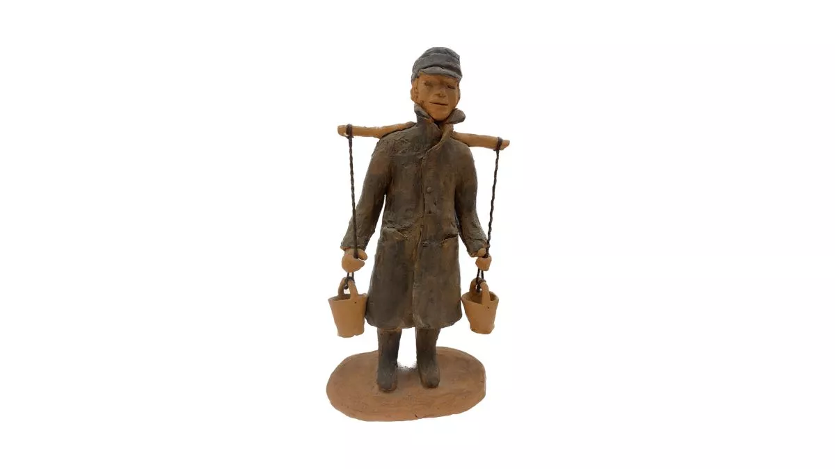 Clay sculpture of a man wearing a long dark grey coat, with dark grey boots and cap. Work uniform including a white shirt, dark grey vest, pants, boots and cap. A long stick is balanced horizontally on his back with two pieces of twine connected to clay buckets tied at both ends. The man’s hands are loosely griped on both pieces of twine, just above the dangling buckets.