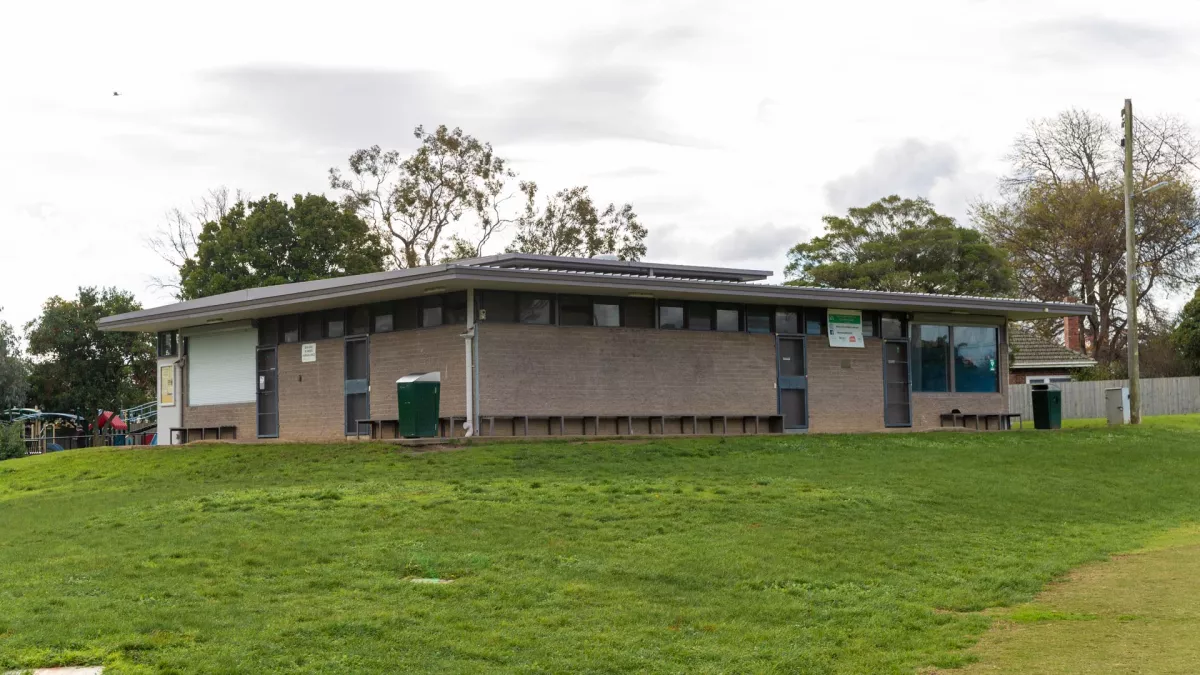 Single-storey rectangular grey brick building with four black doors and a flat roof, on the edge of a sportsground. There are windows above the brickwork. There are several large trees in the background. 