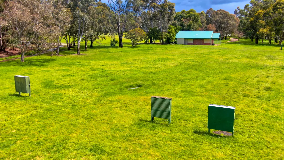 Large rectangular grass area surrounded by medium-sized trees. There are 3 archery targets in the foreground and a small single-storey building at the far end.