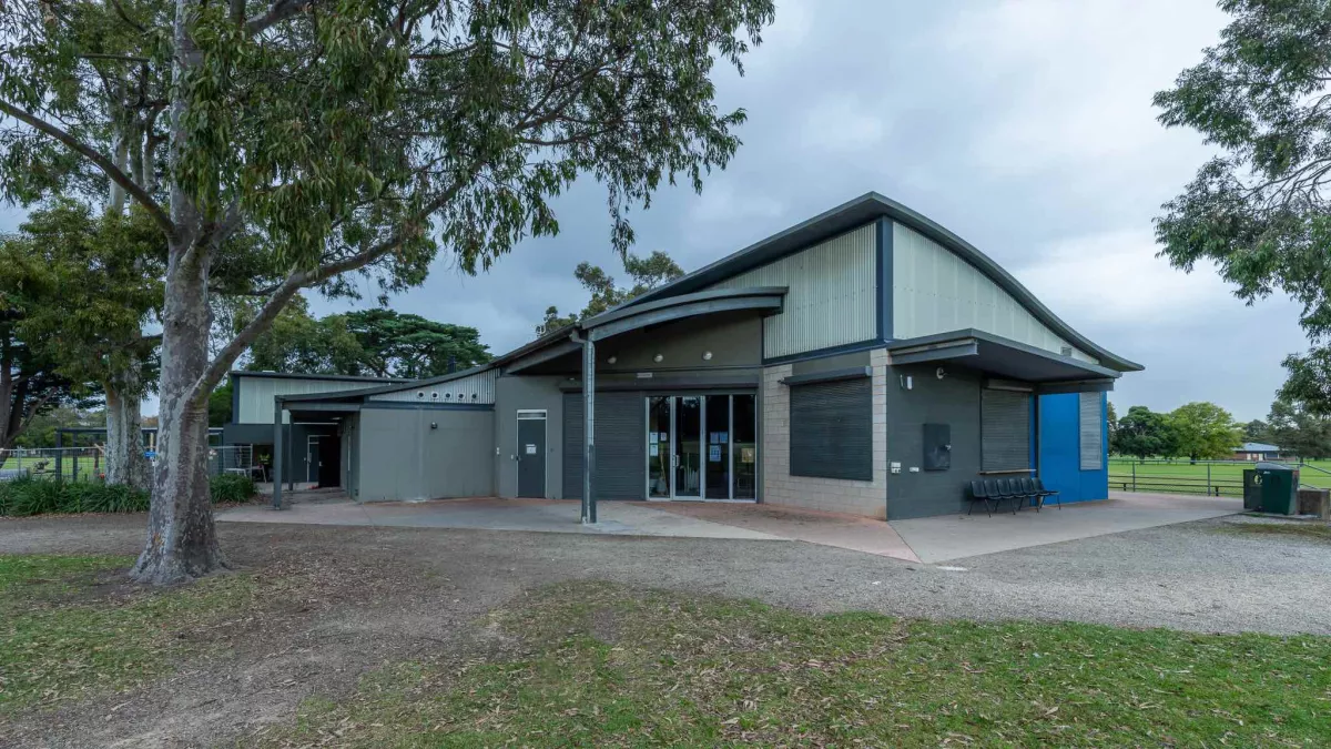 Single-storey grey and white building with glass doors and a triangular corrugated roof. There is a tall, mature tree in the foreground and part of a grass field in the background.