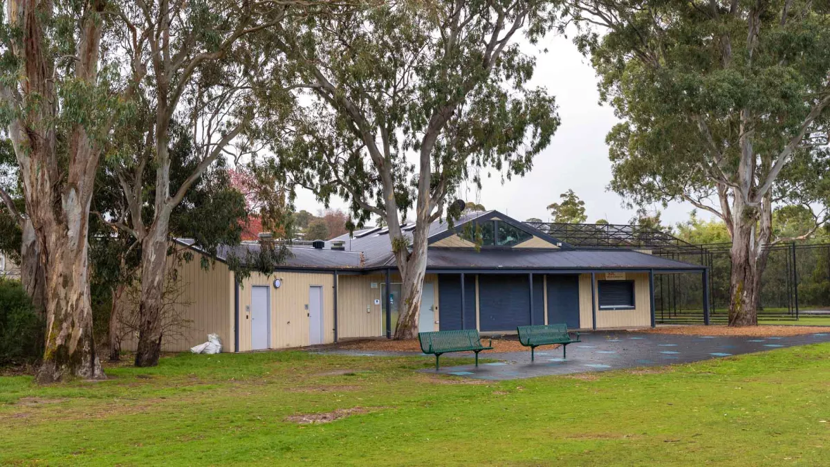 Single-storey dark blue and cream white weatherboard building on the edge of a grass field. There are four mature trees and two park benches at the front of the building. Cricket nets are visible in the background.
