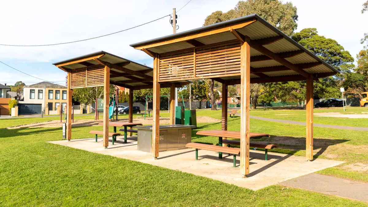 Barbecue area in a park, with 2 sets of tables and chairs and 2 wooden shelter structures with diagonal roofing.