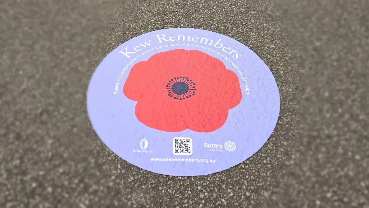 A floor decal on pavement, displaying a red poppy on a purple background with the heading 'Kew Remembers' and a qr code