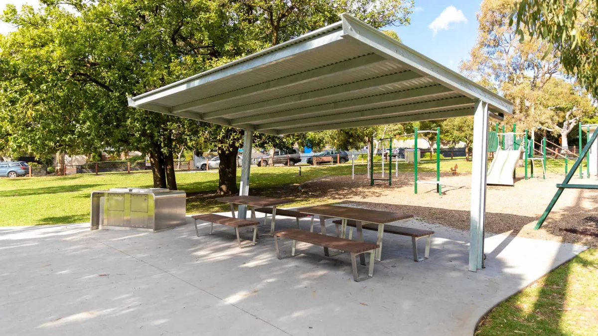 Two sets of tables and bench chairs under a small corrugated iron cover. There is a barbecue to the left and a playground in the background.