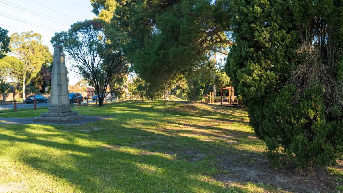 Small grass area with tall concrete memorial structure to the left and tree shadows throughout. There is a thick tree and bare dirt patches to the near right. A road and cars are visible to the left.
