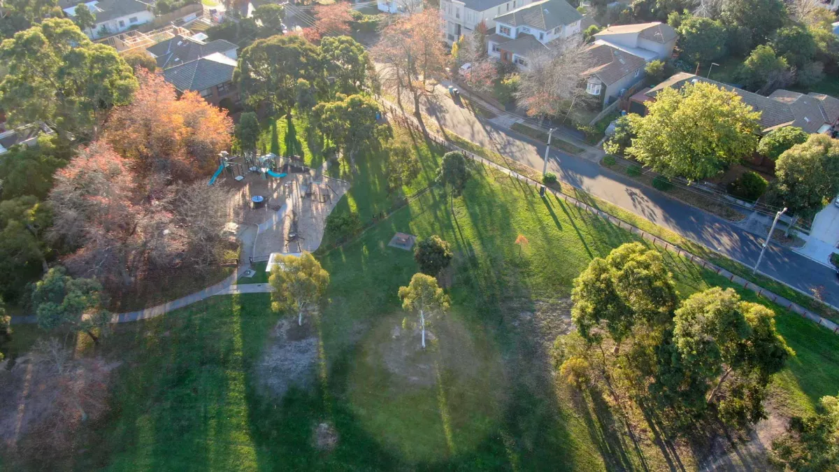 Aerial view of small grass area with a playground and scattered small and medium trees casting long shadows. There is a road and houses at the top right.