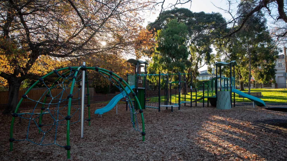 Playground in shadows with large climbing feature, two slides and a dome-shaped feature. There are tall trees and large houses in the background, with sunlight coming through.