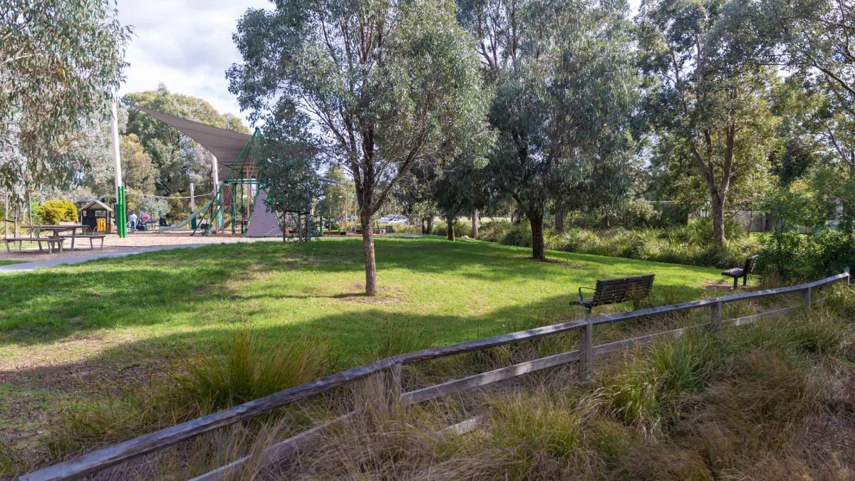 A small grass area separated from a grass shrub area by a wooden fence. The grass area has a couple of trees and wooden seats. in the background is a playground covered by a shade sail and a wooden picnic table. 