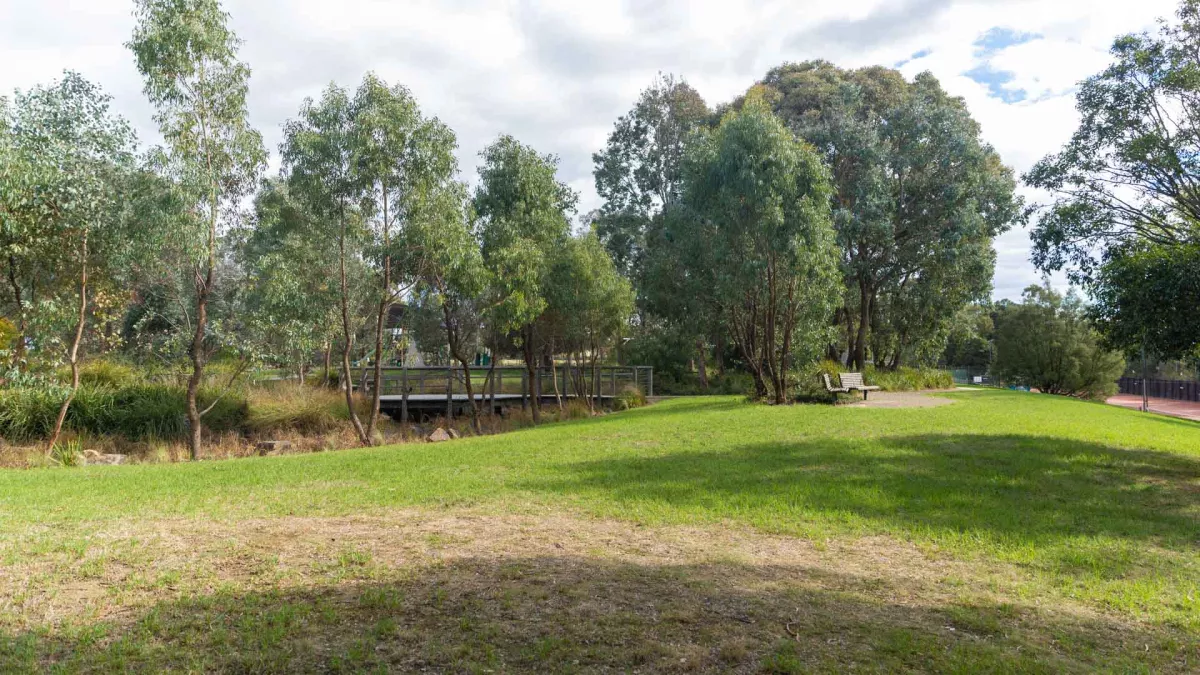 Small grass area with a tree-lined creek and foot bridge to the far left. Two park benches are to the far right. There are shadows and a more bare patch of turf in the foreground.