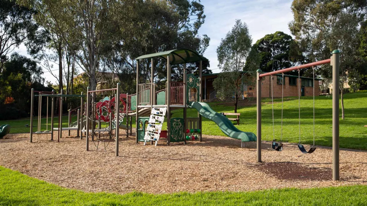 Playground with a large climbing feature, slide, swings and smaller features. There are tall trees and part of a building in the background.