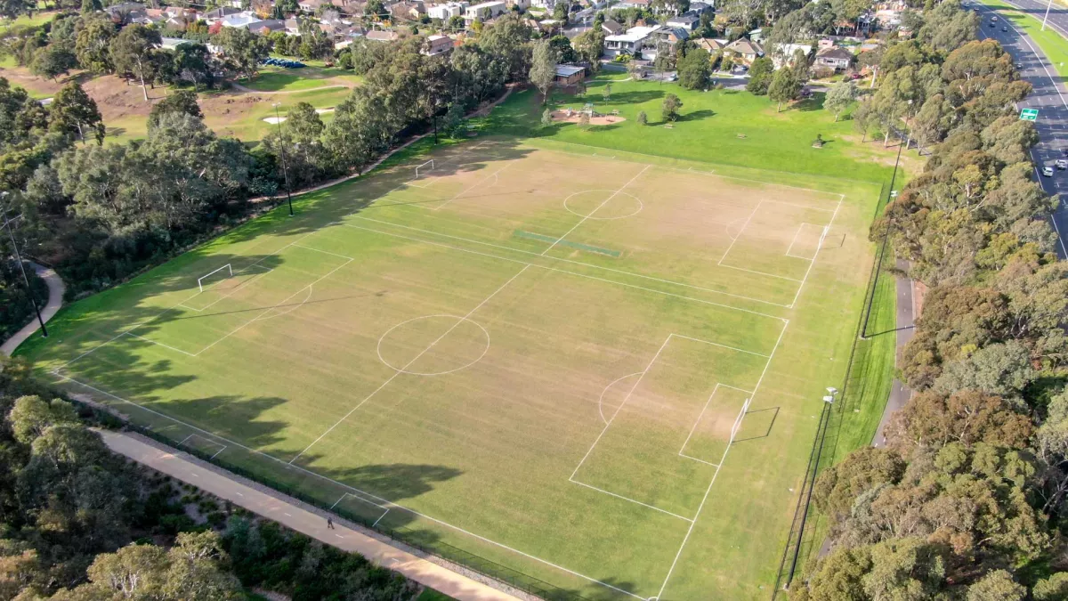 Aerial view of rectangular sports ground with two soccer pitches side-by-side. There is a thick layer of trees to the left and right and houses in the distance.
