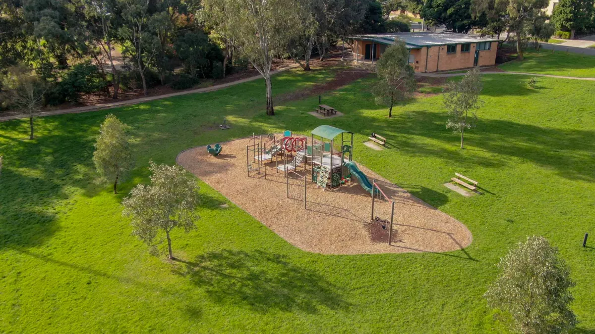 An overhead image of a playground with a mulch floor. It is surrounded by a large grass area. A walking path runs along the edge separating the park from a tree area. A brick building is also in the background. 