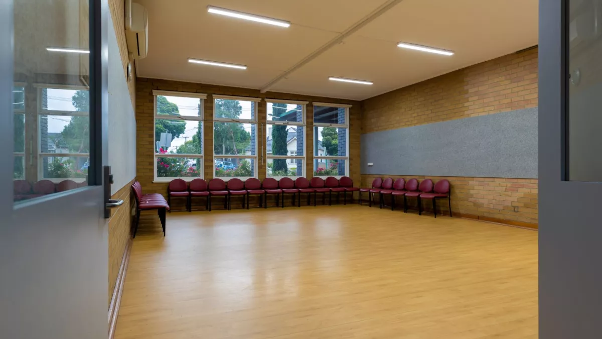 A small empty room with a wooden fall. Chairs lines the back wall of the room. Large windows look out towards the tress outside. 