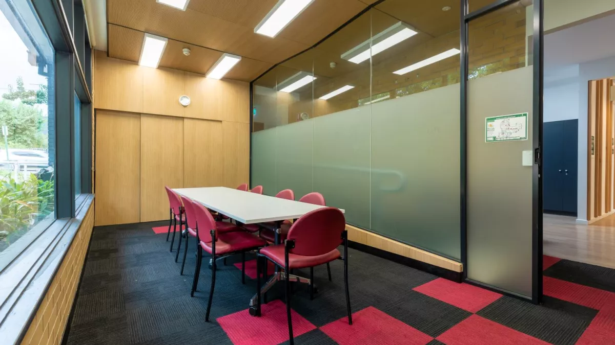 A small room with a rectangular table surrounded by red chairs. A large window is on the left wall, and a obscured glass wall on the other side of the room
