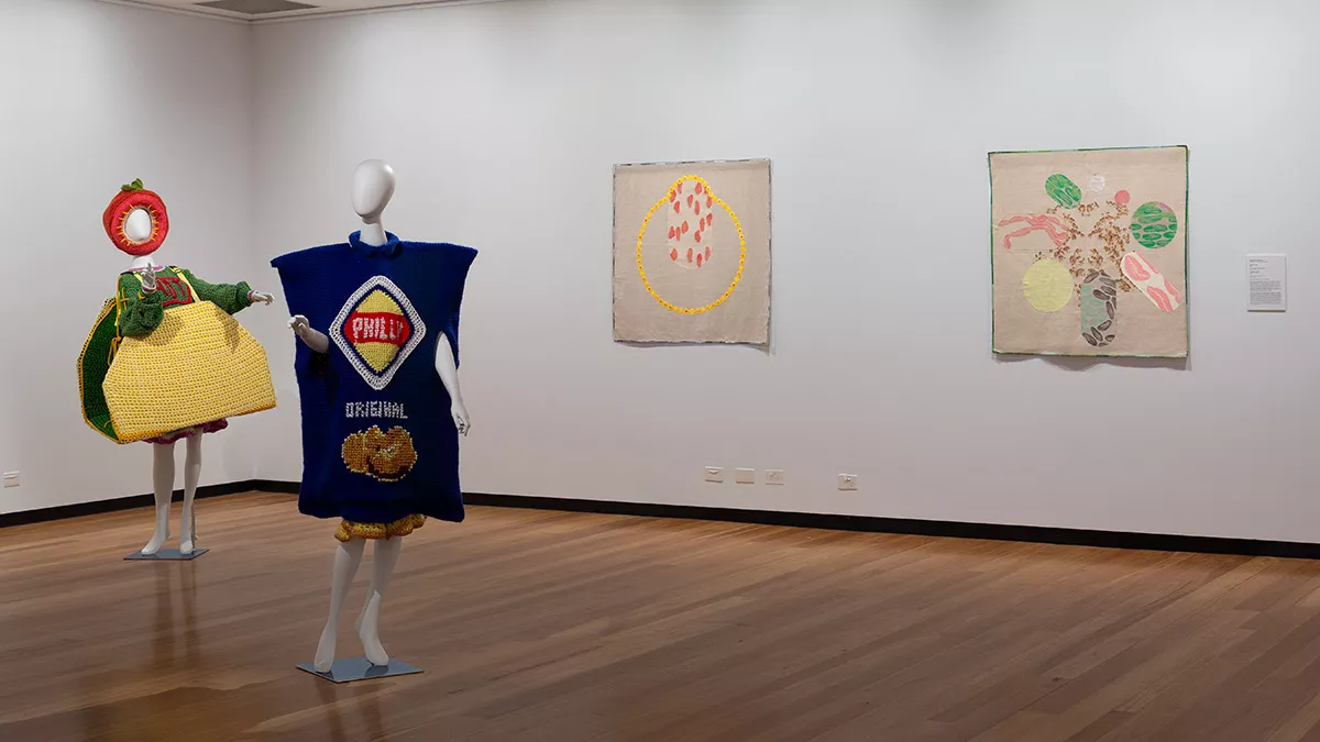Two mannequins wearing colorful outfits stand in front of a gallery wall with three colorful artworks.