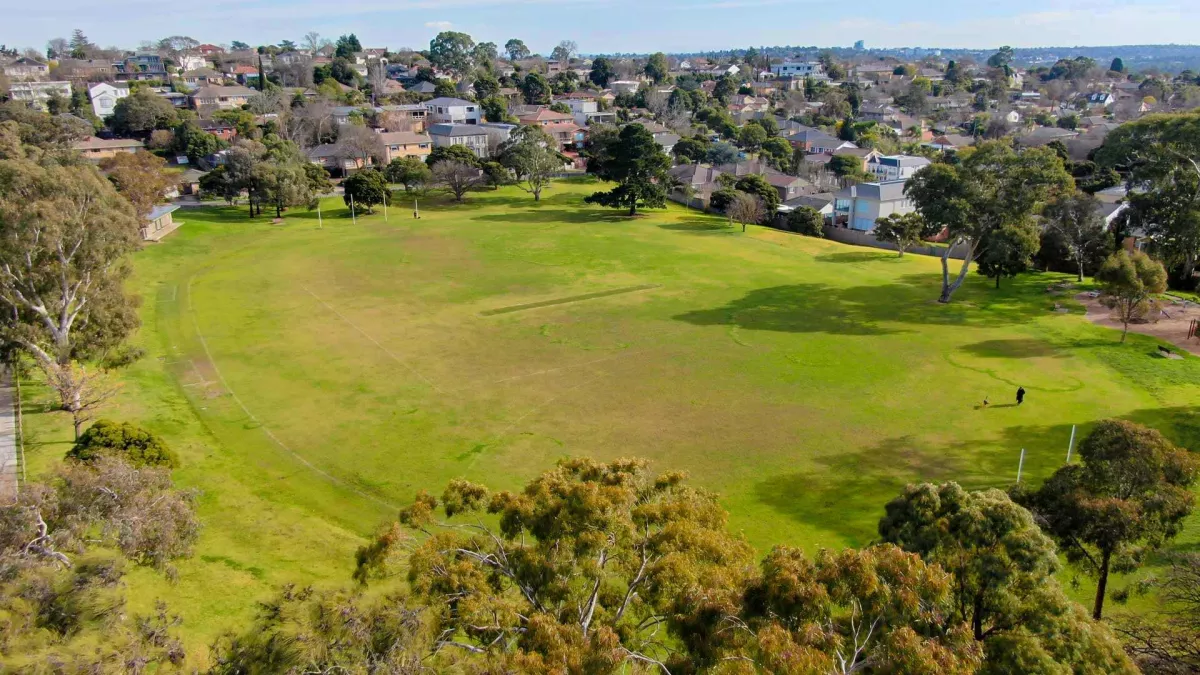 An above ground view of a green park. it is surrounded by trees and houses can be seen in the background