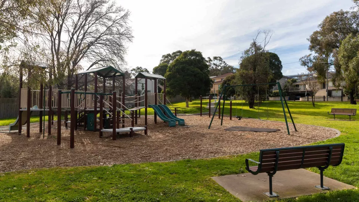 A kids playground featuring a climbing frame, a slide and a swing set on a mulch floor. Wooden benches face towards the playground