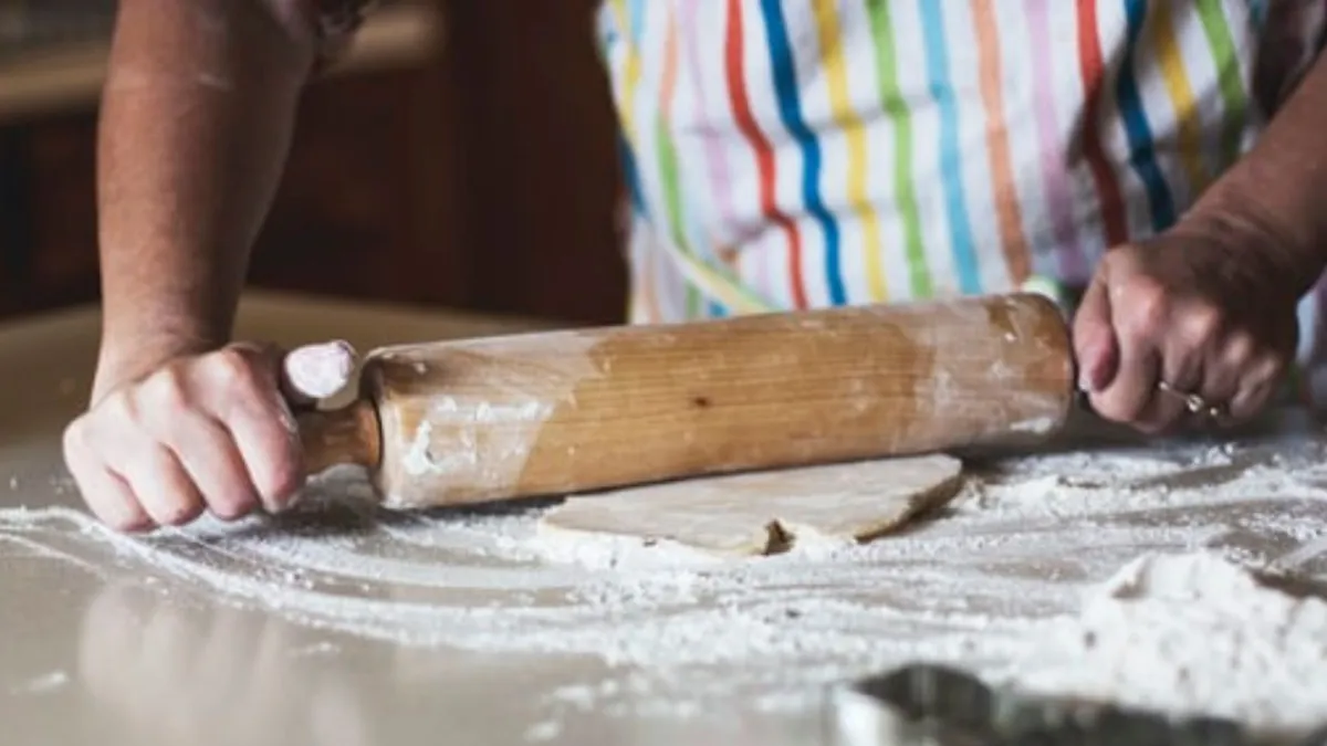 photo of a person rolling dough with a rolling pin. They are wearing a striped apron and there is lots of flour on the bench.