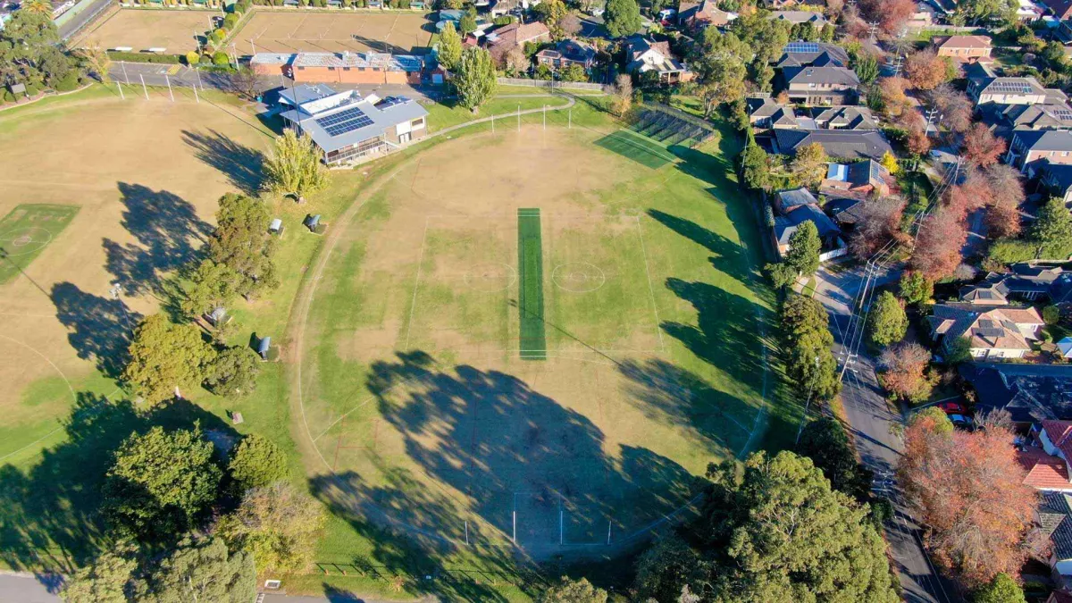 Aerial view of an oval sportsgrouind. A green cricket pitch is in the centre. The oval is surrounded by houses and trees. 