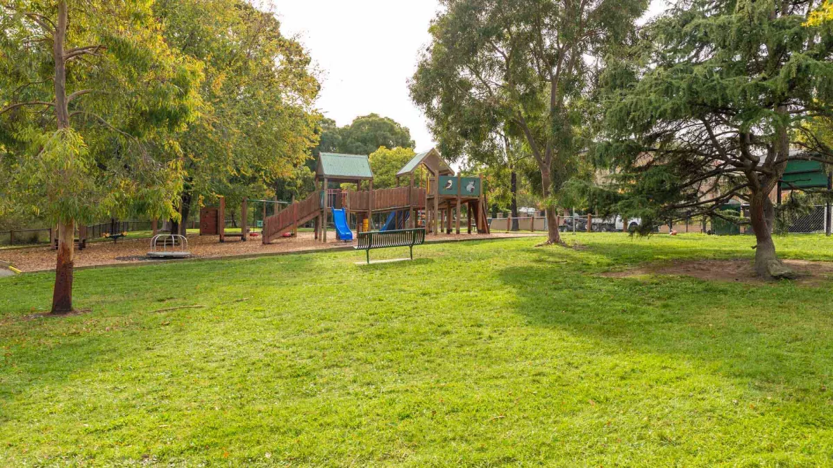 Tall trees and a metal park bench surround a wooden playground. 