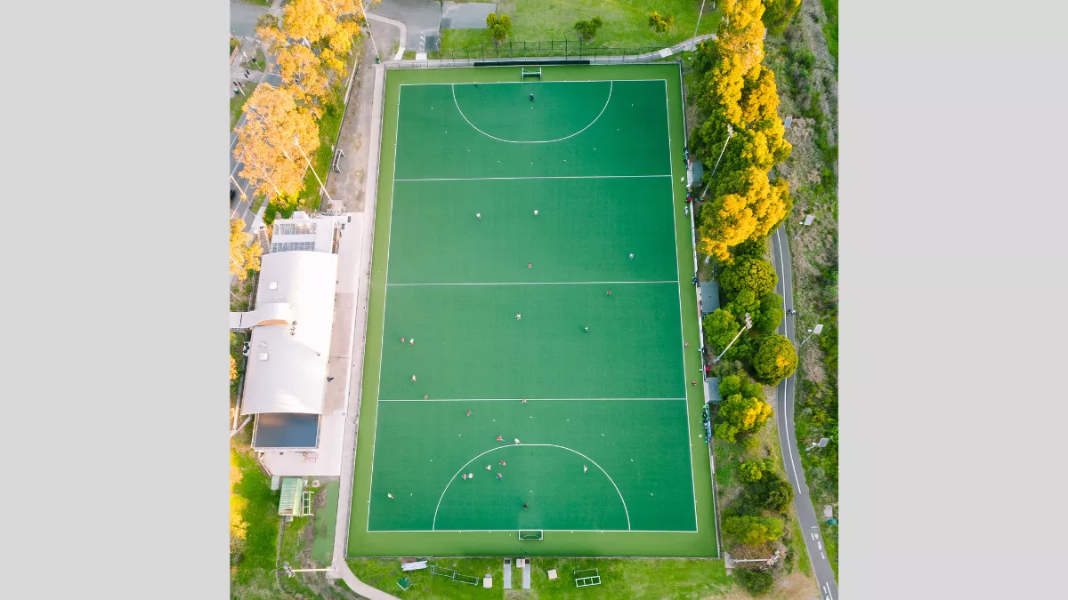 An aerial view of a rectangular sports field, presumably a hockey field, featuring a synthetic green playing surface marked with white lines. The field is surrounded by a boundary with a buffer zone of artificial turf or grass. Several small figures, likely players, are scattered across the field engaged in various activities. On the left side of the image, adjacent to the field, there is a white-roofed building, possibly a clubhouse or facility associated with the sports field. 