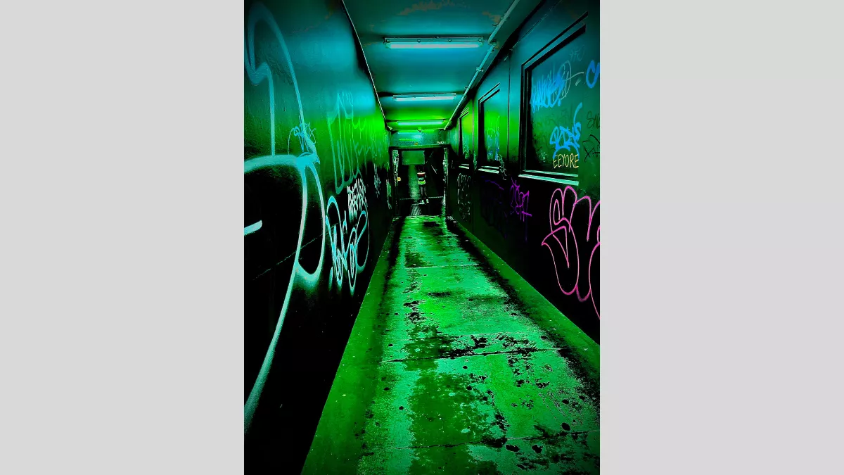 A narrow, indoor corridor with walls covered in vibrant graffiti. The corridor is bathed in a greenish light, creating a surreal and edgy atmosphere. The graffiti art features various tags and designs, adding to the urban aesthetic.