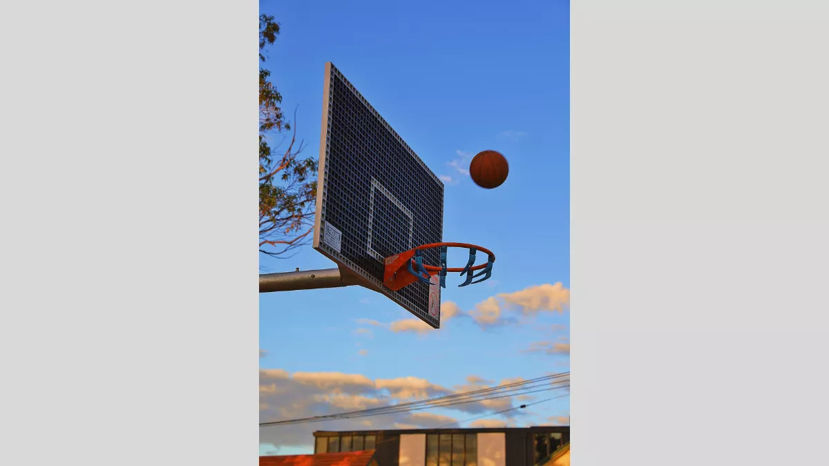 A dynamic shot of a basketball in mid-air approaching a hoop. The scene is set against a clear blue sky with a few scattered clouds. The background includes tree branches and rooftops, adding to the outdoor setting.