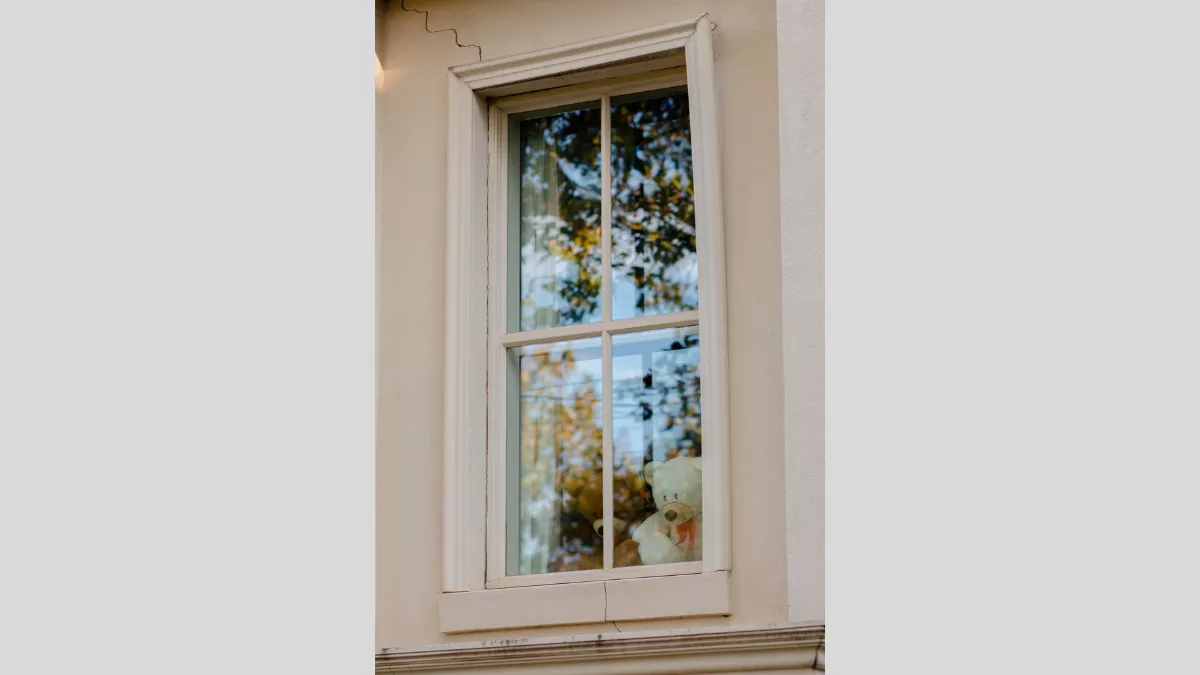 Photograph of two teddy bears in a long rectangular window. The teddy on the left is brown and the one on the right is white with a red bowtie around its neck. The façade of the house is painted a light coral colour, and the outside of the window reflects the trees outside. 