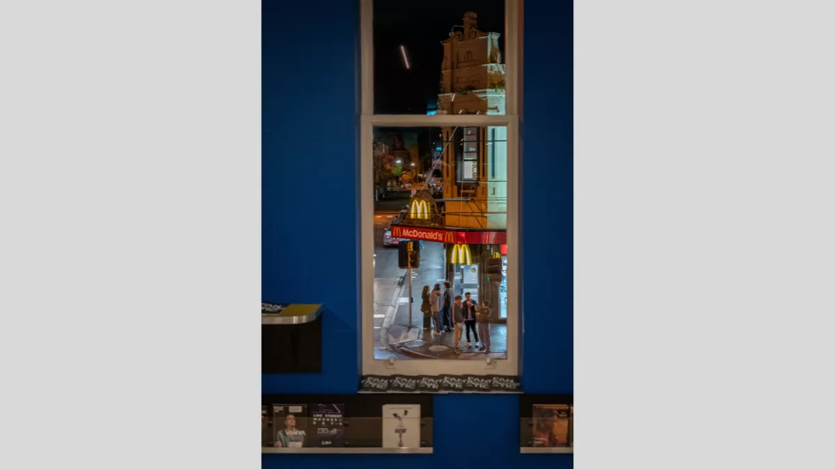 A nighttime urban scene viewed through a window. The image captures a McDonald's restaurant sign illuminated in the night, with people gathered on the street below. The scene is framed by the window, offering a glimpse of the city life outside