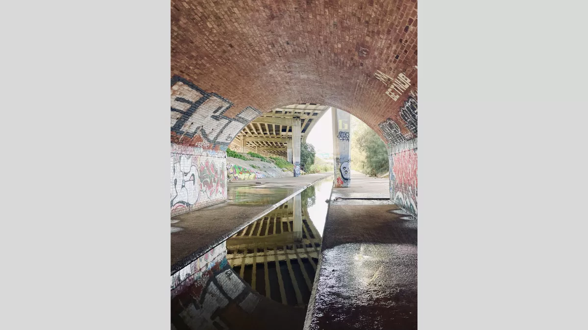 A photograph taken under a bridge with graffiti-covered walls. The scene shows a puddle reflecting the bridge's arch and the graffiti, creating a symmetrical visual effect. The background features another bridge and a path extending into the distance.
