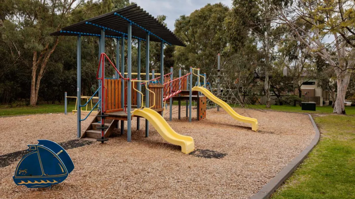 A playground with a bark flow. It has 2 slides and a rocking seat. A barbecue can also been seen.