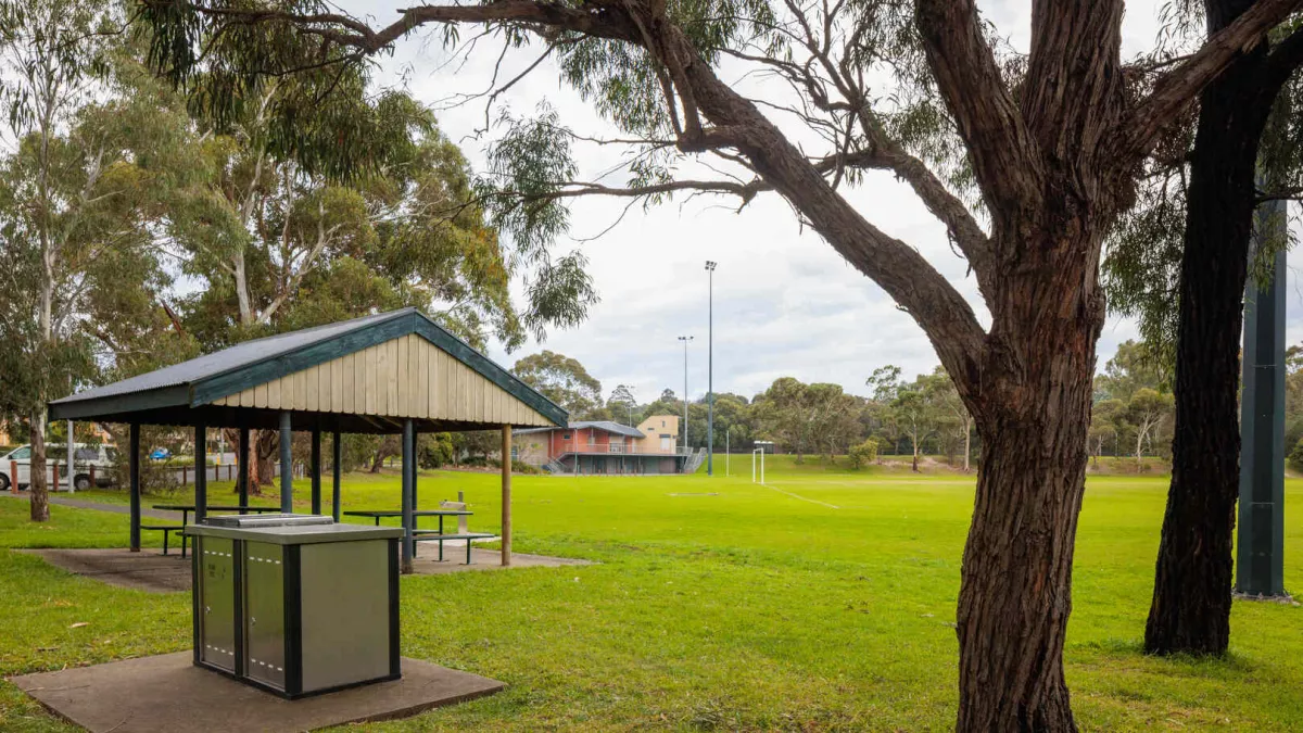 A park area with a barbecue and an undercover seating area. A sportsfield is in the background.