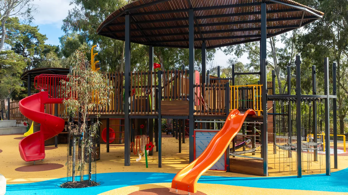 a vibrant playground with a substantial wooden play structure at its heart. The multi-tiered design incorporates a slide, climbing wall, and monkey bars, among other features. The surrounding area is shaded by several trees, and the ground is cushioned with a rubberized safety surface. The playground is both engaging and secure, offering children an enjoyable play environment.
