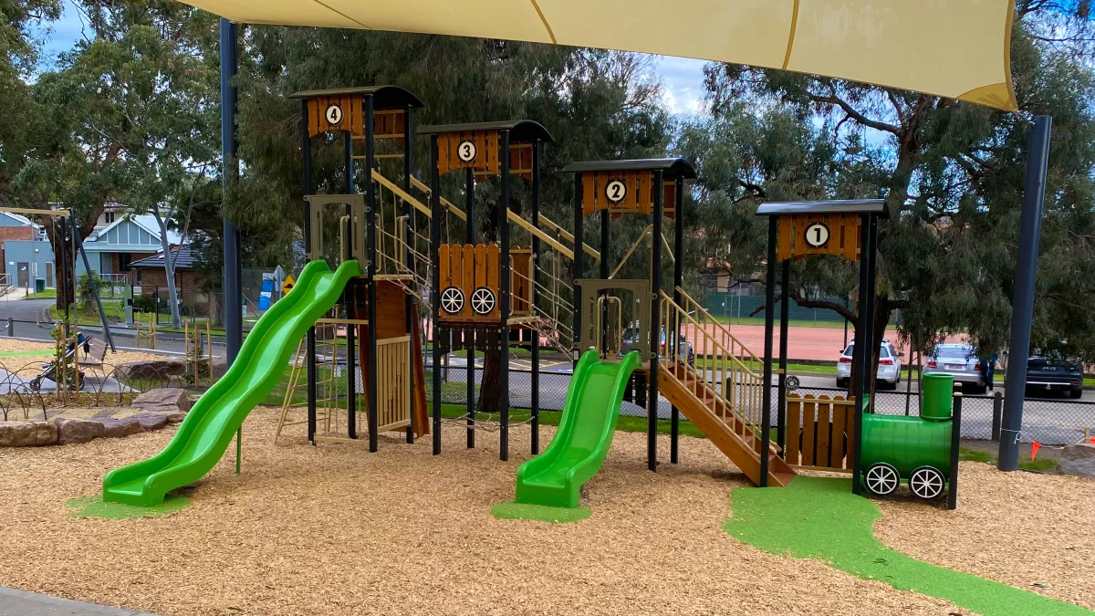 A children's playground has a timber climbing frame with green slides beneath a shade sail