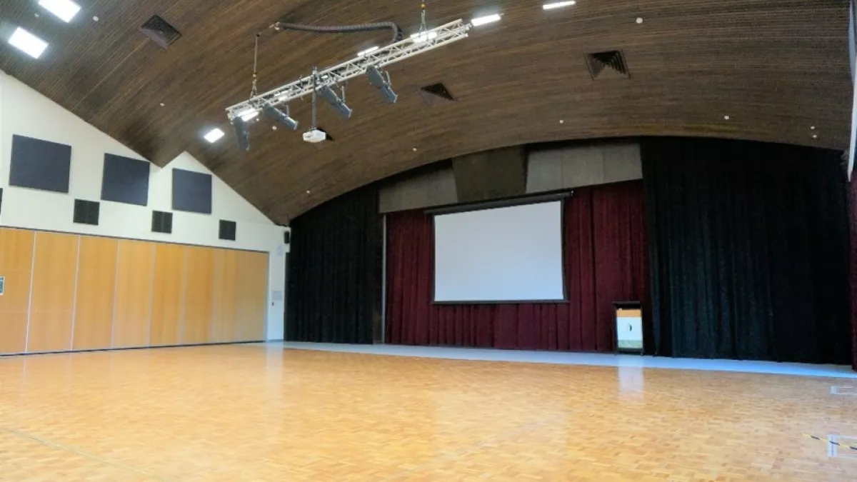 A large empty room with a wooden floor, panelled ceiling. At the back of the room is a projection screen and red curtains
