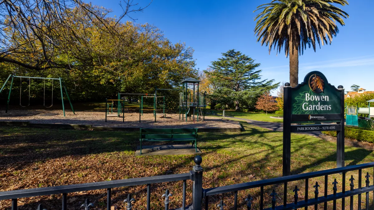 A green children playground surrounded by mulch. There is a black iron fence surround the playground with a sign that says Bowen Gardens. 