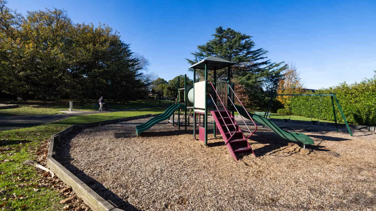 A close up of a children's playground with a tambark floor featuring 2 slides. Behind the playground is a walking path. 