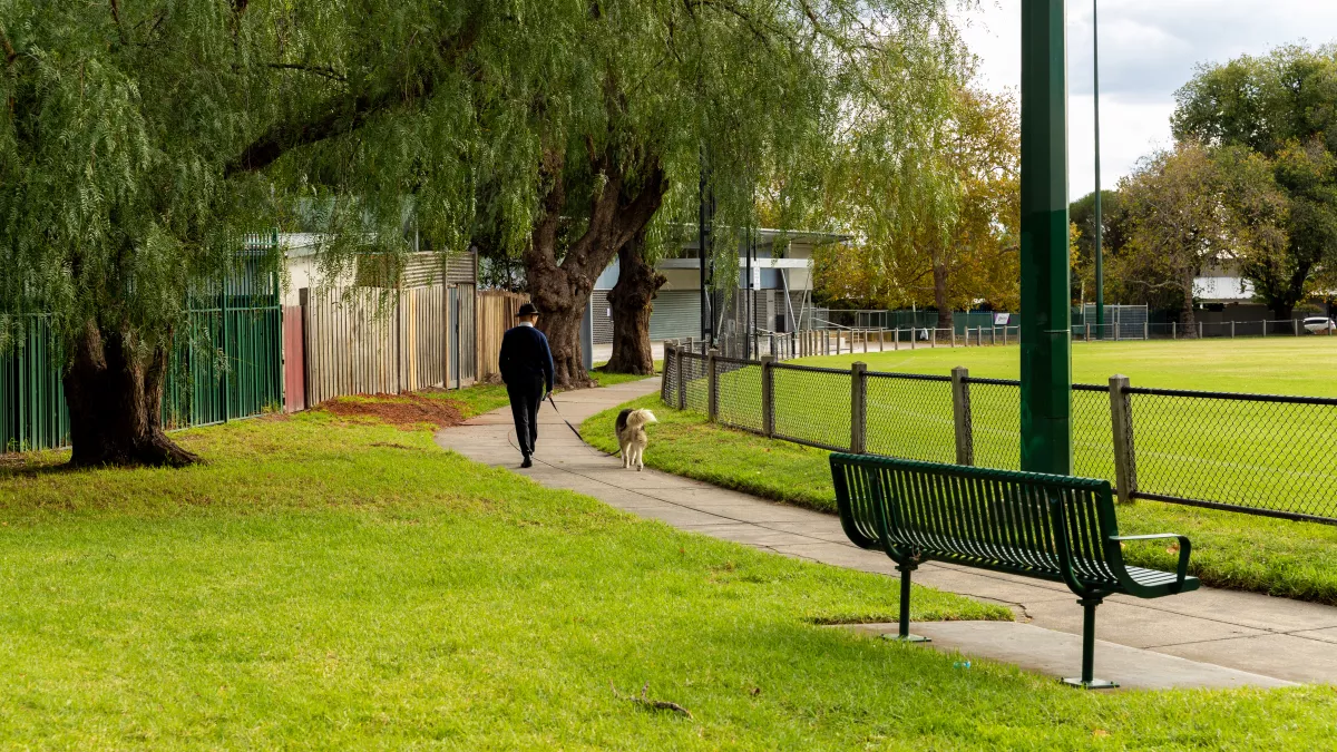 A person walks a dog on a path around a grassy sportsground