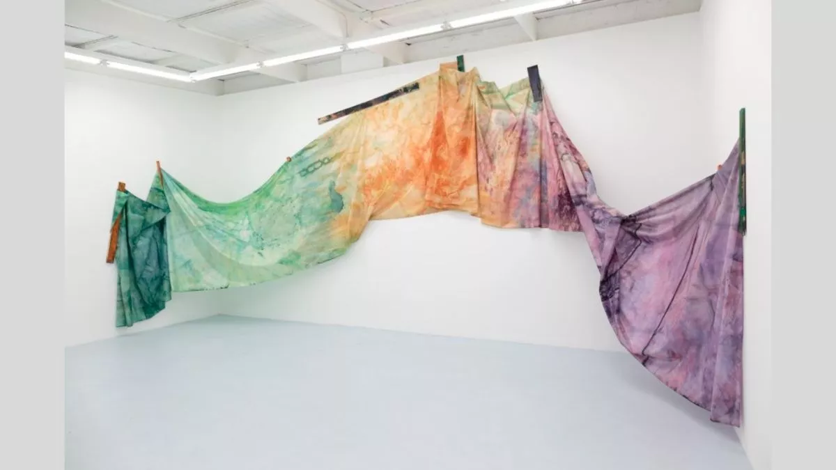 This image shows an installation artwork featuring a large, dyed fabric panel. The fabric is suspended and draped across the corner of a white gallery space, attached to wooden supports that are fixed to the walls. The fabric displays variations in colours that shift from green, to orange and purple, creating a gradient-like effect, and is arranged in a flowing, wave-like formation. The installation interacts with the surrounding architecture, with the fabric appearing to move between tension and looseness.