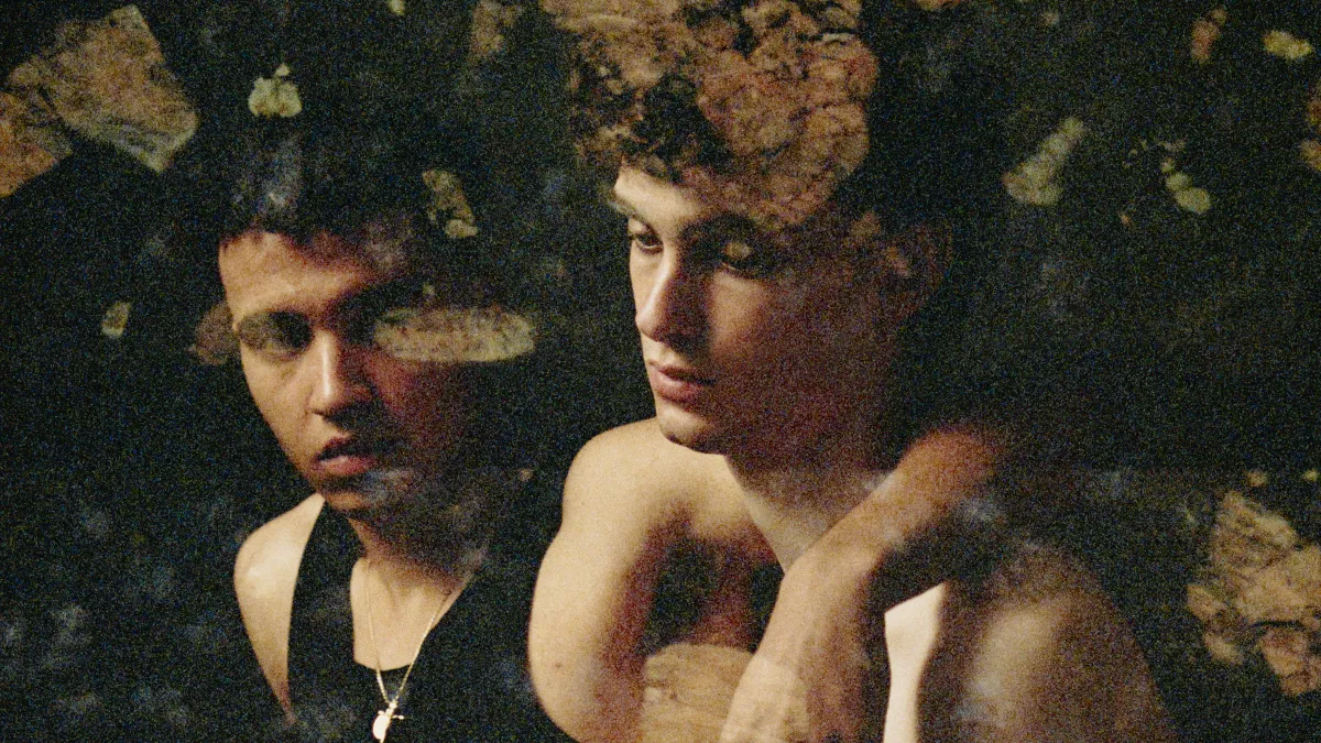 A photograph of two male figures sitting side by side against a black background. One has his arm over the shoulder of the other and is looking at him. The other is looking downwards and is holding a pale pink rose. Patches of semi-transparent brown botanical pattern cover the photograph. 