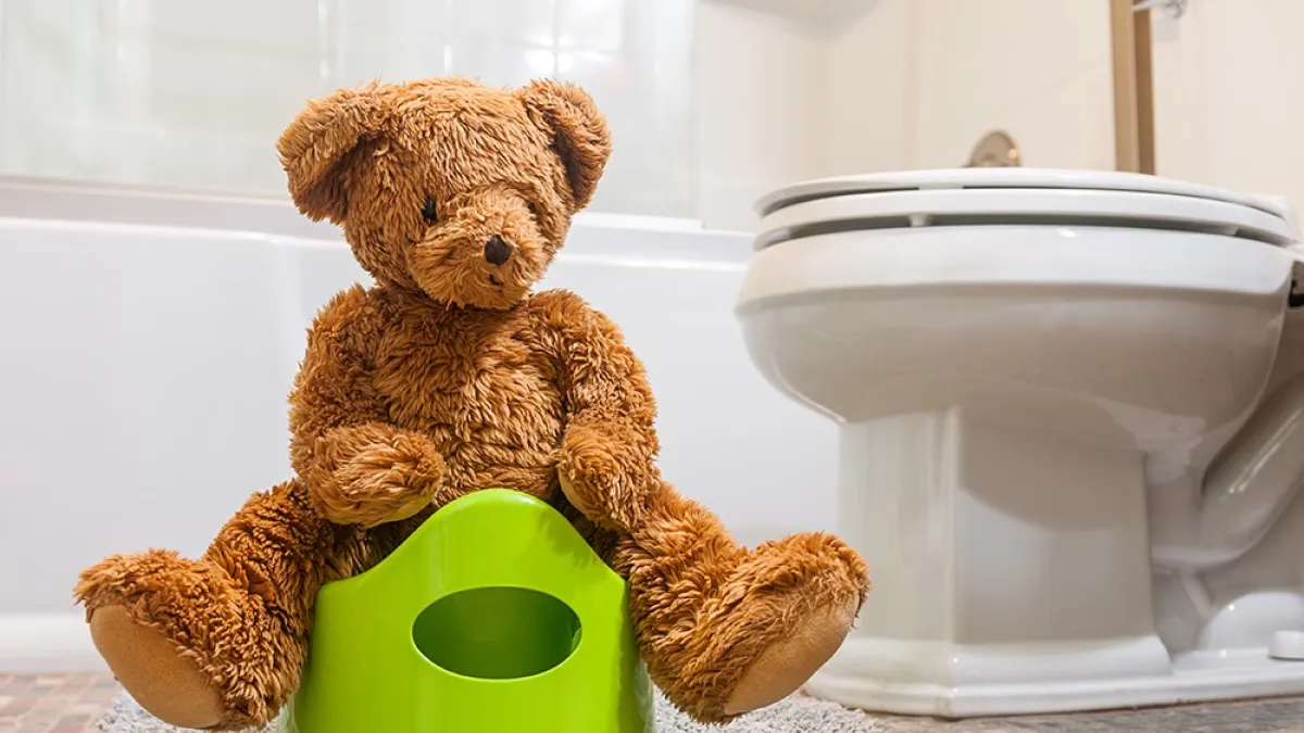 A toy teddy bear sits on a green potty, in a bathroom.