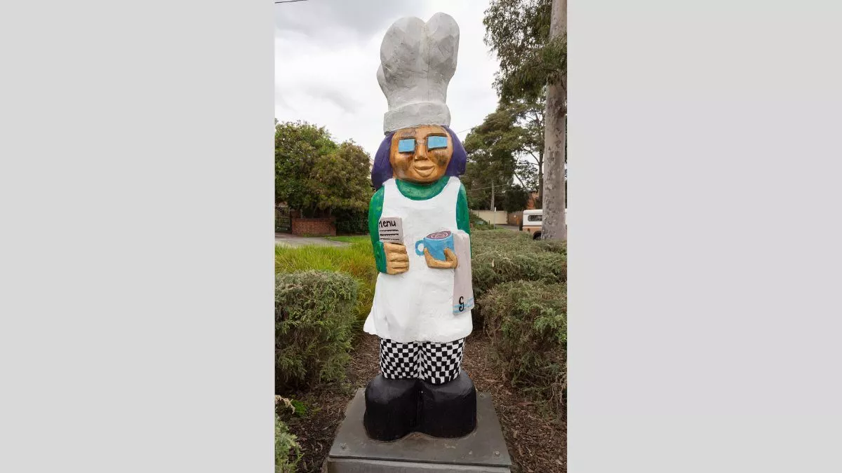 A carved wooden figure of a baker wearing a large white chef’s hat, an apron and black and white checkered pants. The baker holds a newspaper and a coffee, with a tea towel slung over their arm.