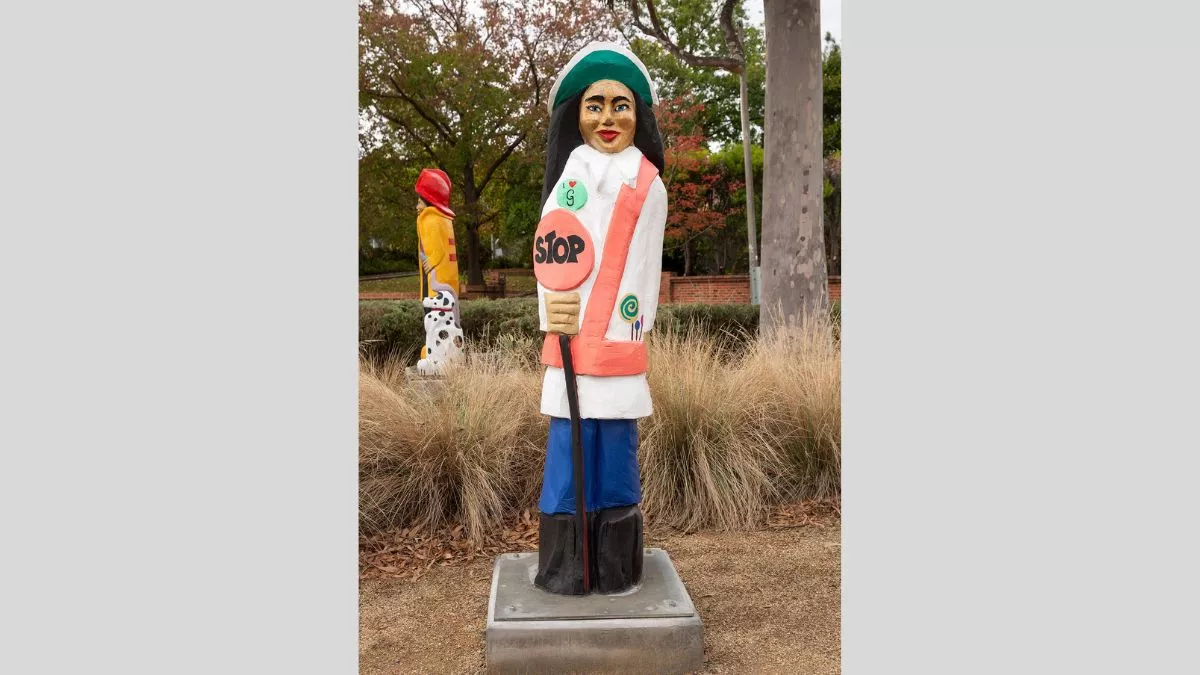 A carved wooden figure of a school crossing supervisor. The lady wears a long white coat and green hat. Some lollipops are peeking out of her pocket.
