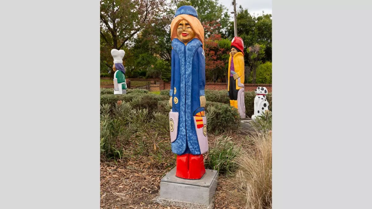 A carved wooden figure of a glamorous woman with a long blue coat. She has blond hair, bright red boots and carries two shopping bags.